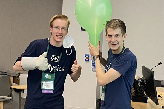 Two students in GW Physics t-shirts at a conference demonstrating with a large green lightbulb-like object